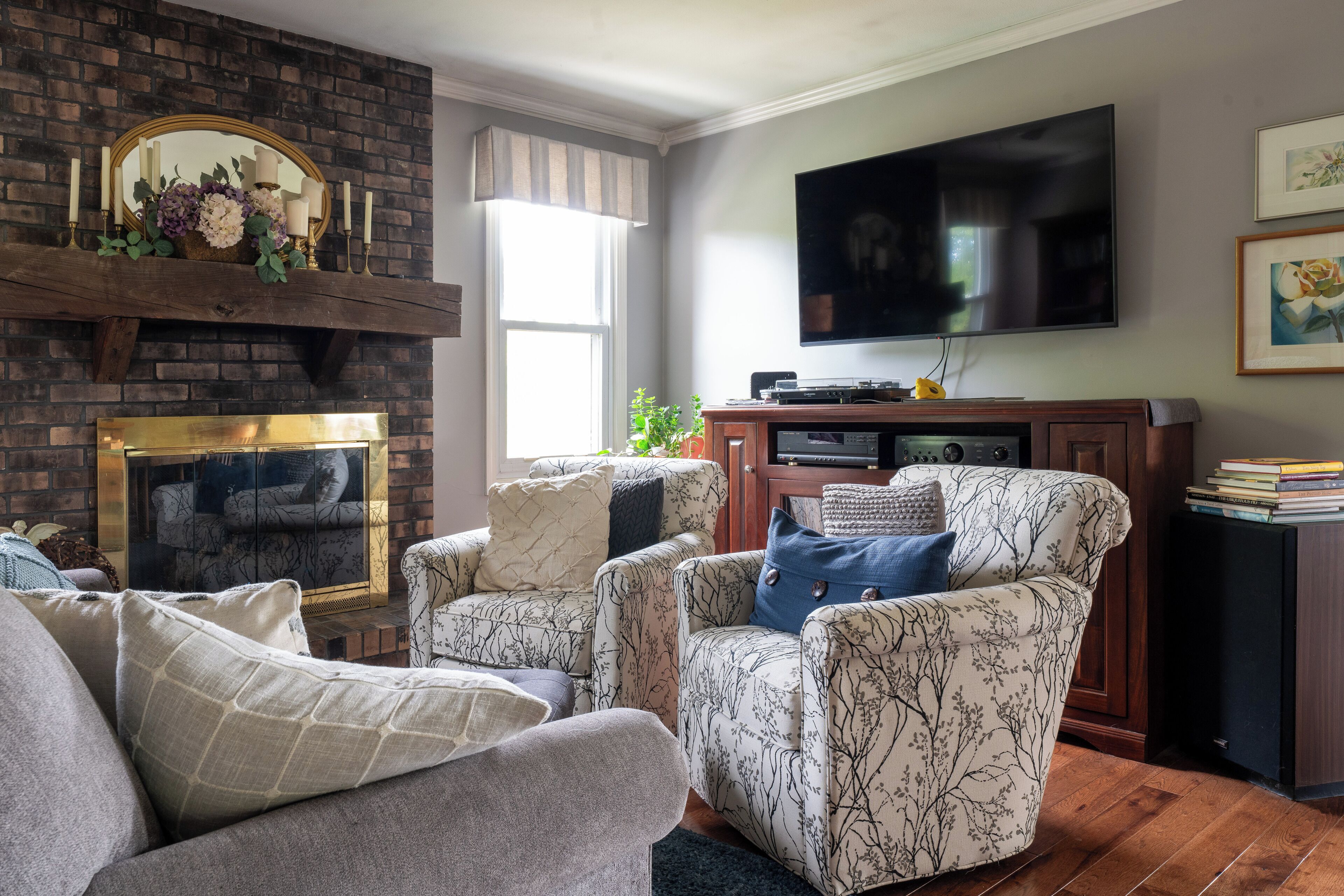 A living room with a brick fireplace, two patterned armchairs, a sofa, a flat-screen TV, and a bookshelf.
