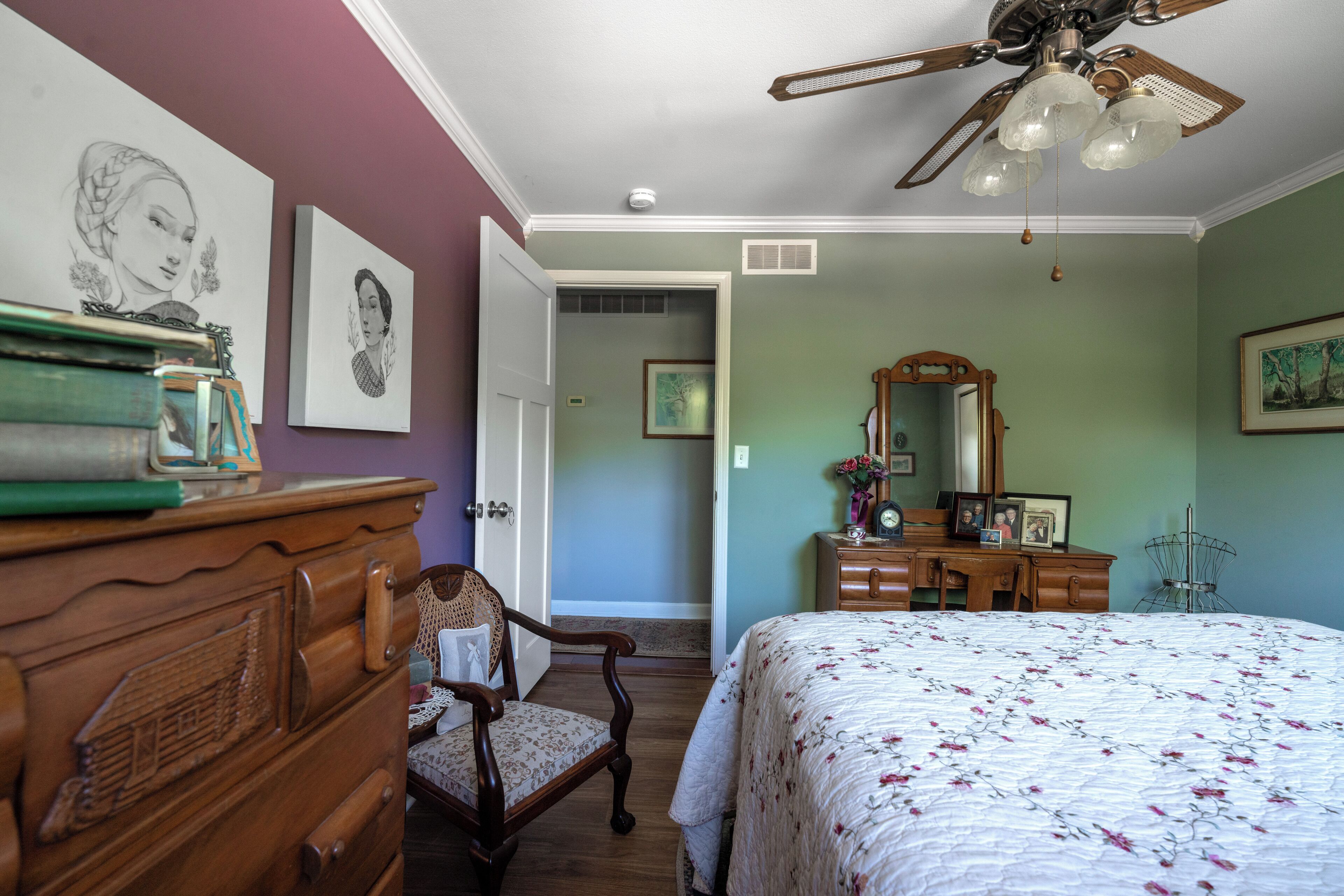 A bedroom with a wooden dresser, a chair, a bed with a floral bedspread, a ceiling fan, and artwork on the walls.