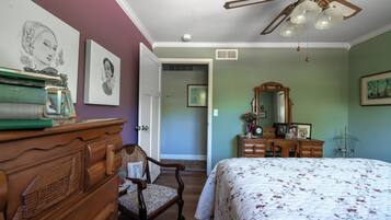 A bedroom with a wooden dresser, a chair, a bed with a floral bedspread, a ceiling fan, and artwork on the walls.
