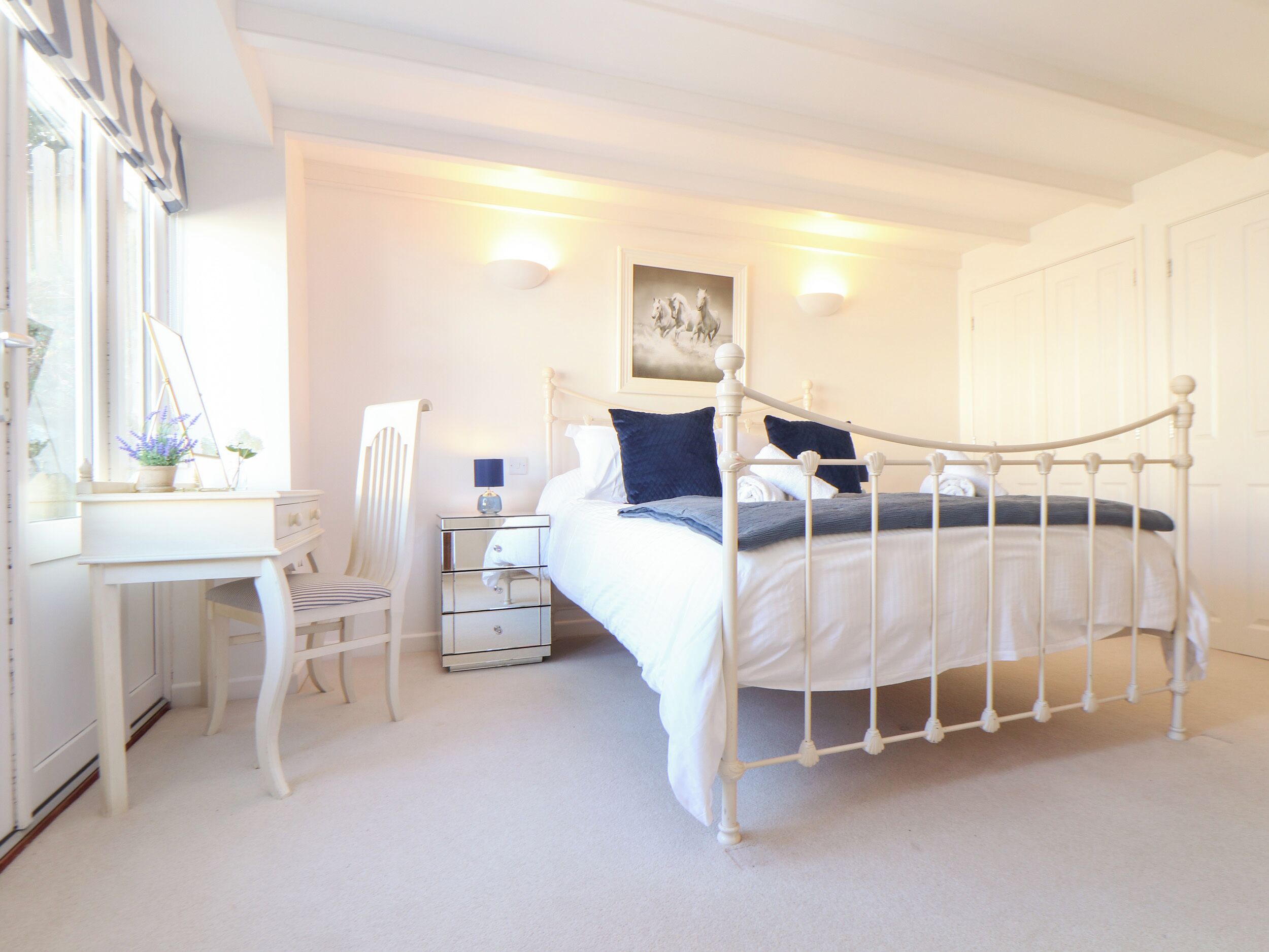 A bedroom with a white metal bed frame, a bedside table with a lamp, a chair, and a desk with a mirror and a plant.