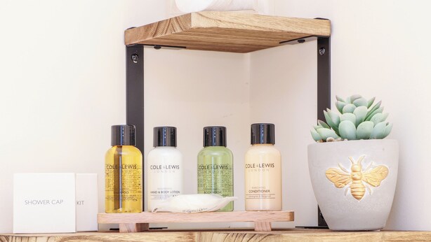 Una estantería con cuatro botellas de productos Cole & Lewis London, un gorro de ducha y una planta en maceta con diseño de abeja.