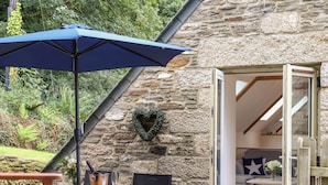 Une terrasse extérieure avec une table, des chaises et un parasol bleu. On peut voir un mur de pierre orné d’une guirlande en forme de cœur.