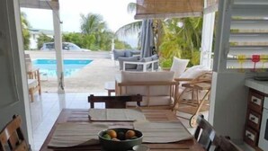 Comedor con mesa de madera dispuesta para cuatro, un frutero y vistas a una piscina y zona de estar exterior.