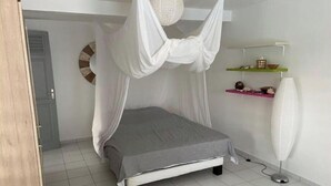 Un dormitorio con una cama bajo dosel blanco, una mesita de noche con lámpara y un pequeño estante con objetos decorativos.