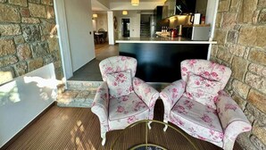 A cozy seating area with two floral armchairs and a small table, a stone wall, and a view into a modern kitchen and dining area. - Panoramic Villa with jacuzzi (Bodrum)