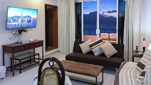 Ein Wohnzimmer mit einer Couch, einem Fernseher, einem kleinen Tisch und einem Sessel. Durch große Fenster ist der Meerblick zu sehen.