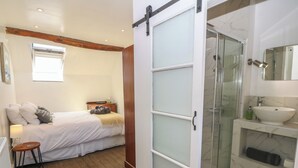 Ein Schlafzimmer mit Bett, Nachttisch, Lampe und einem kleinen Hocker. Daneben befindet sich ein Badezimmer mit Dusche, Waschbecken und Spiegel.