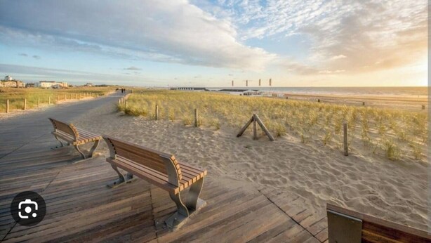 En træpromenade der fører til en strand med klitter og et fjerntliggende havnemole.