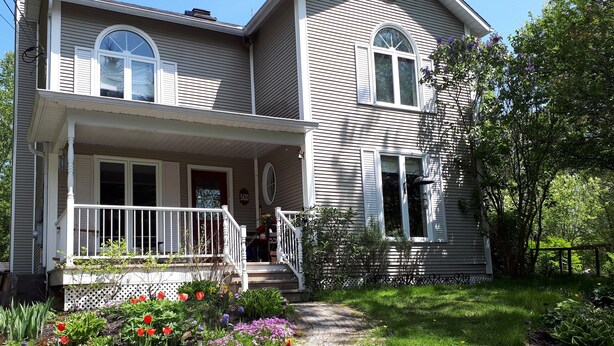 A two-story house with a front porch, a mailbox, and a garden with various plants and flowers. - Intimité et Confort, Plusieurs Foyers, à Proximité D'activités et Sherbrooke (Sherbrooke)