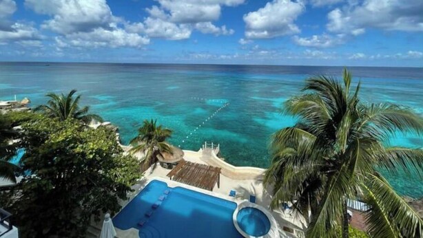 Une piscine d’un complexe hôtelier, avec une eau turquoise et limpide, entourée de palmiers et offrant une vue sur l’océan sous un ciel partiellement nuageux.