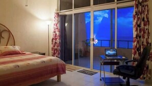 Une chambre d’hôtel avec un lit, un bureau avec un ordinateur portable et une chaise. Il y a une grande fenêtre donnant sur le ciel.