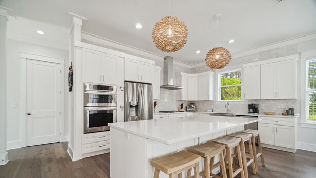 A modern kitchen with white cabinetry, stainless steel appliances, a large island with bar stools, and woven pendant lights. - 30A Beach House - 30A Wonderland by PHG (Panama City Beach)