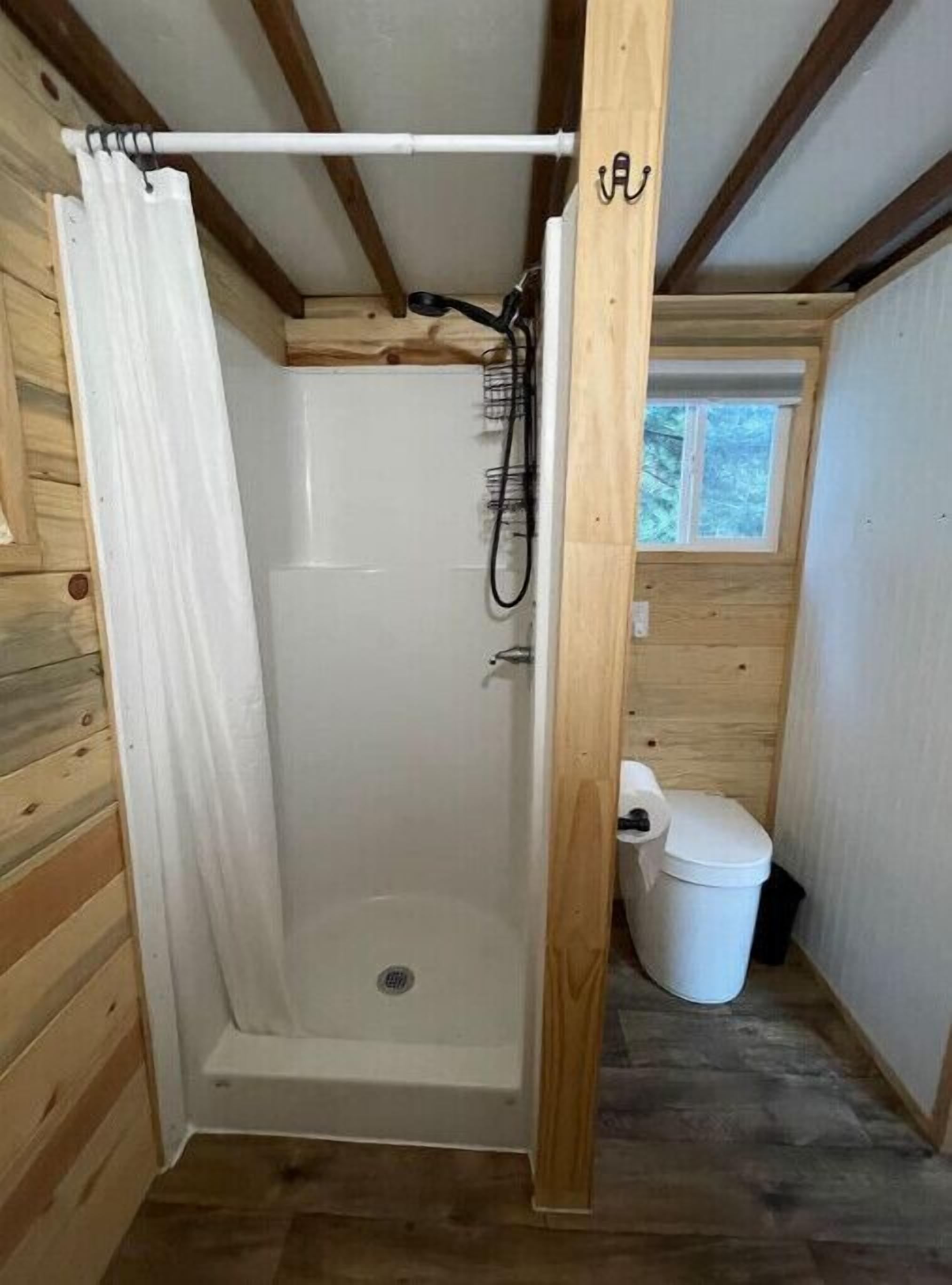 Un petit espace douche fermé, avec un rideau blanc, un plancher en bois et une fenêtre donnant sur les arbres.