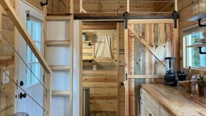 Un interior rústico de madera con una zona de cocina, armarios de madera, un ventilador de techo y una puerta corredera que da a otra habitación.
