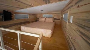 Habitación pequeña con paneles de madera, una cama individual, un ventanal y una barandilla de metal.