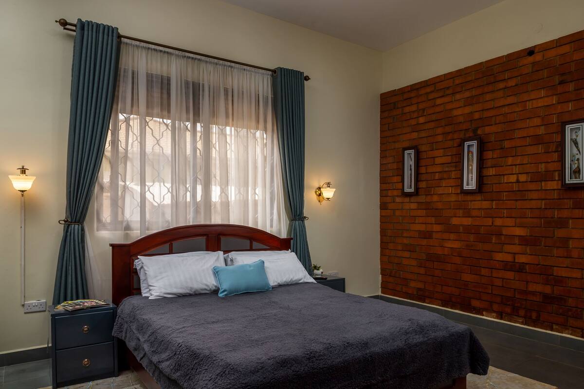 A bedroom with a wooden headboard, a bed with a dark blanket, a nightstand with a lamp, and a brick wall with framed pictures.