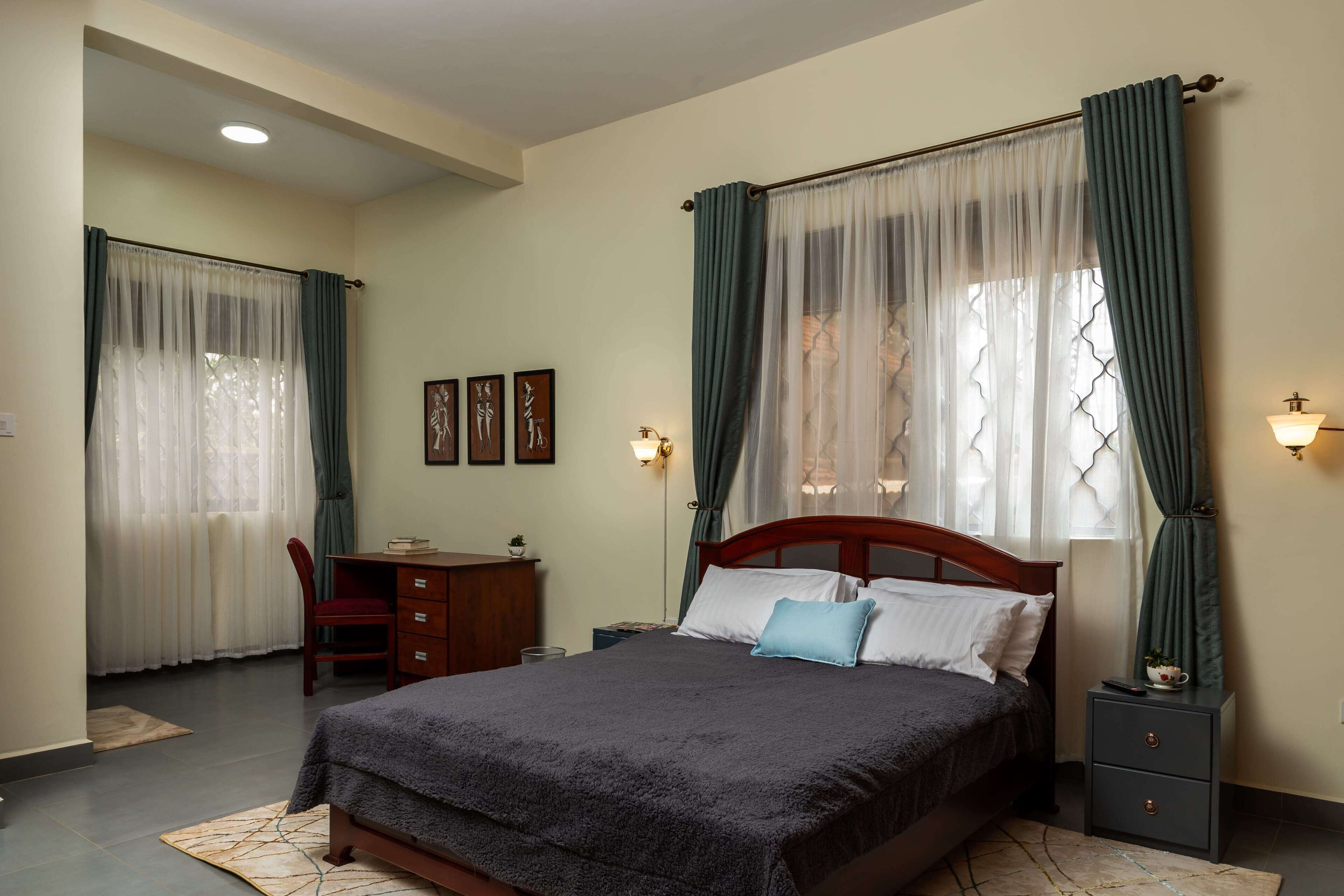 Une chambre à coucher avec un lit, un bureau et une chaise. Il y a des rideaux aux fenêtres et une lampe fixée au mur.