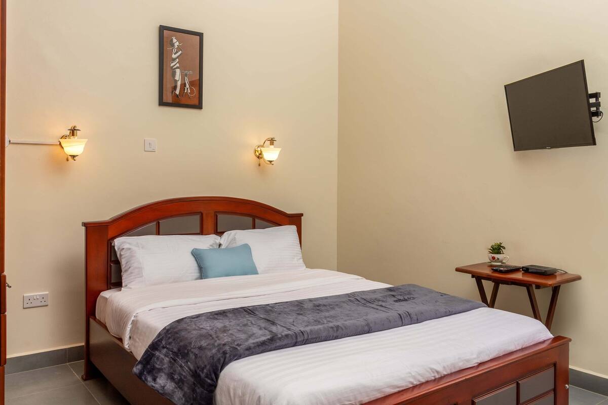 A neatly made bed with a wooden headboard, two wall-mounted lamps, a small table with a plant, and a TV mounted on the wall.