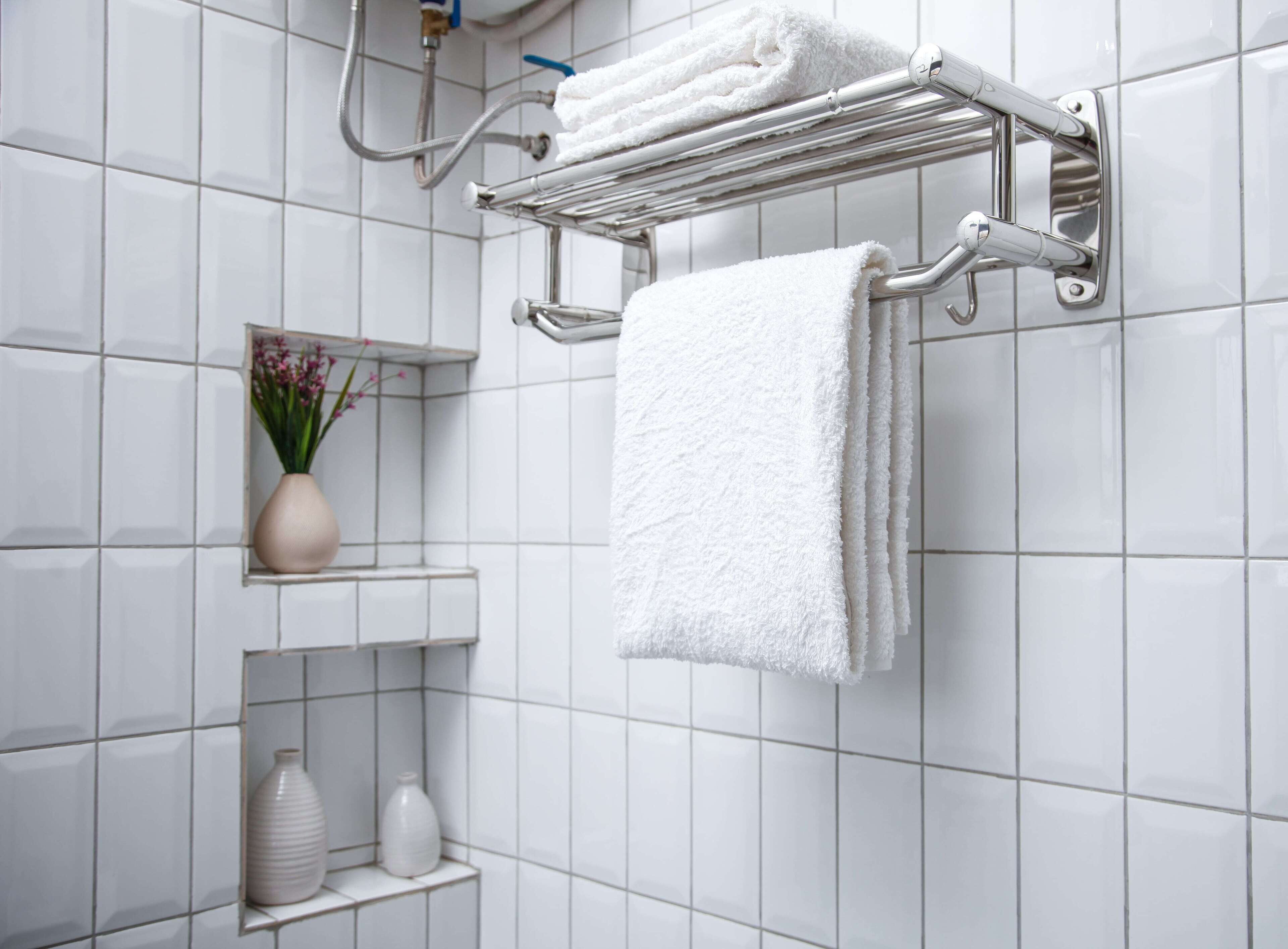 A bathroom with white tiled walls, a towel rack with two towels, and a shelf with a vase and two bottles.
