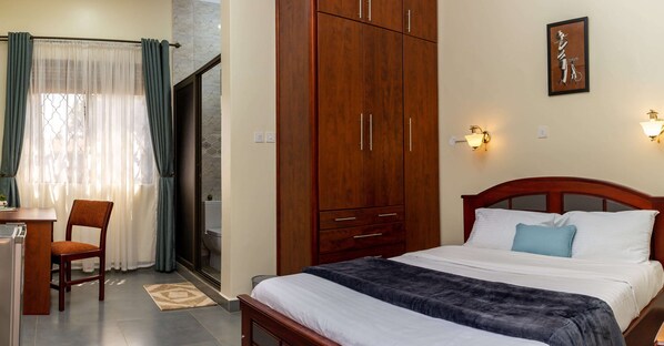 Habitación de hotel con cama, armario de madera, escritorio con silla y baño visible a través de una puerta de cristal.