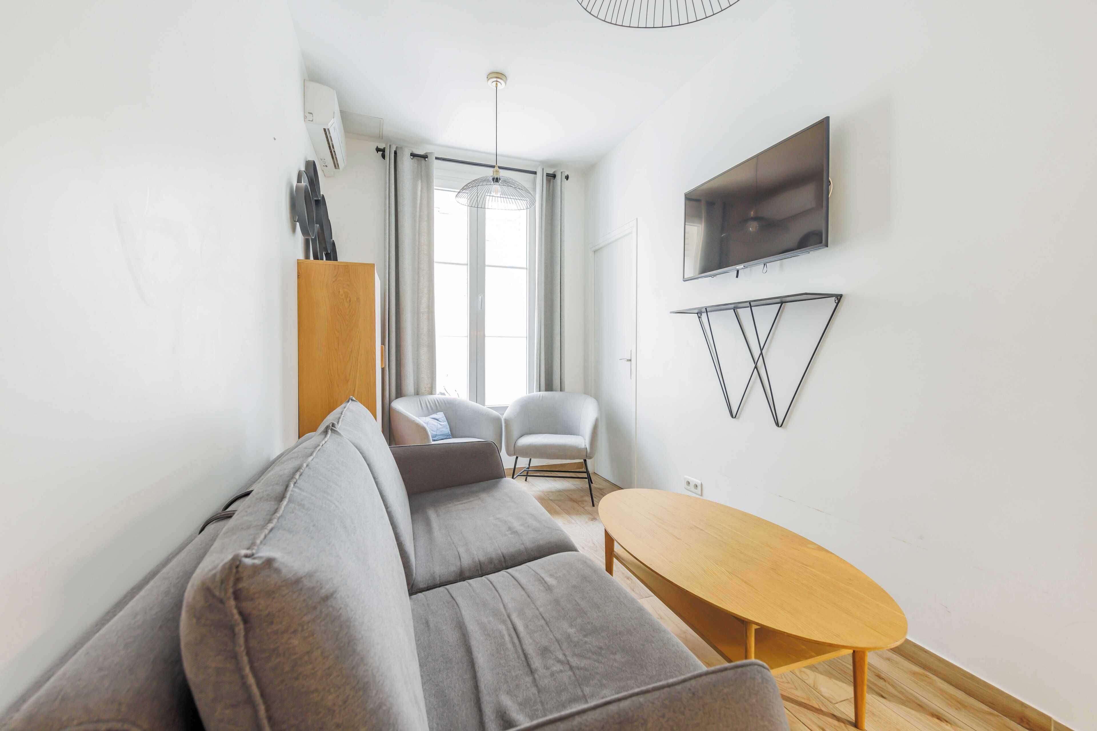 Ein modernes Wohnzimmer mit einem grauen Sofa, zwei Sesseln, einem Holztisch, einem an der Wand montierten Fernseher und einer Pendelleuchte.