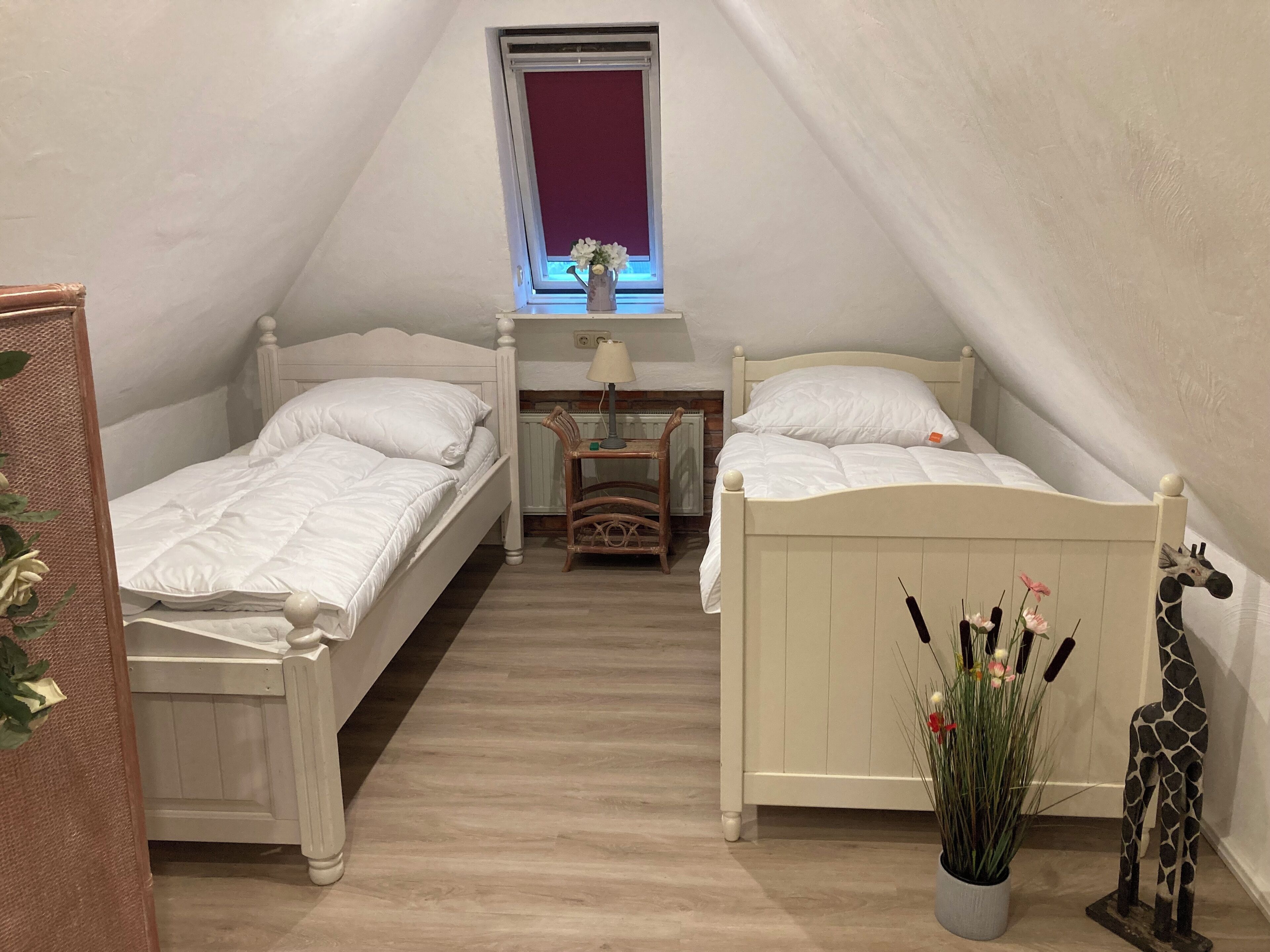 Ein kleines, gemütliches Schlafzimmer im Dachgeschoss mit zwei weißen Betten, einem kleinen Tisch mit Lampe, einem Fenster mit Aussicht und einem Blumentopf mit Giraffendeko.