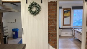 A modern interior with a kitchen area, a white door with a wreath, a brick wall, and a bed in the adjacent room. - Cozy vacation apartment with complete equipment for up to 5 people (Bötersen)