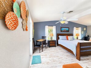 Une chambre à coucher avec un lit, un bureau et une chaise. On y trouve un ventilateur de plafond, une fenêtre avec des rideaux et une porte.