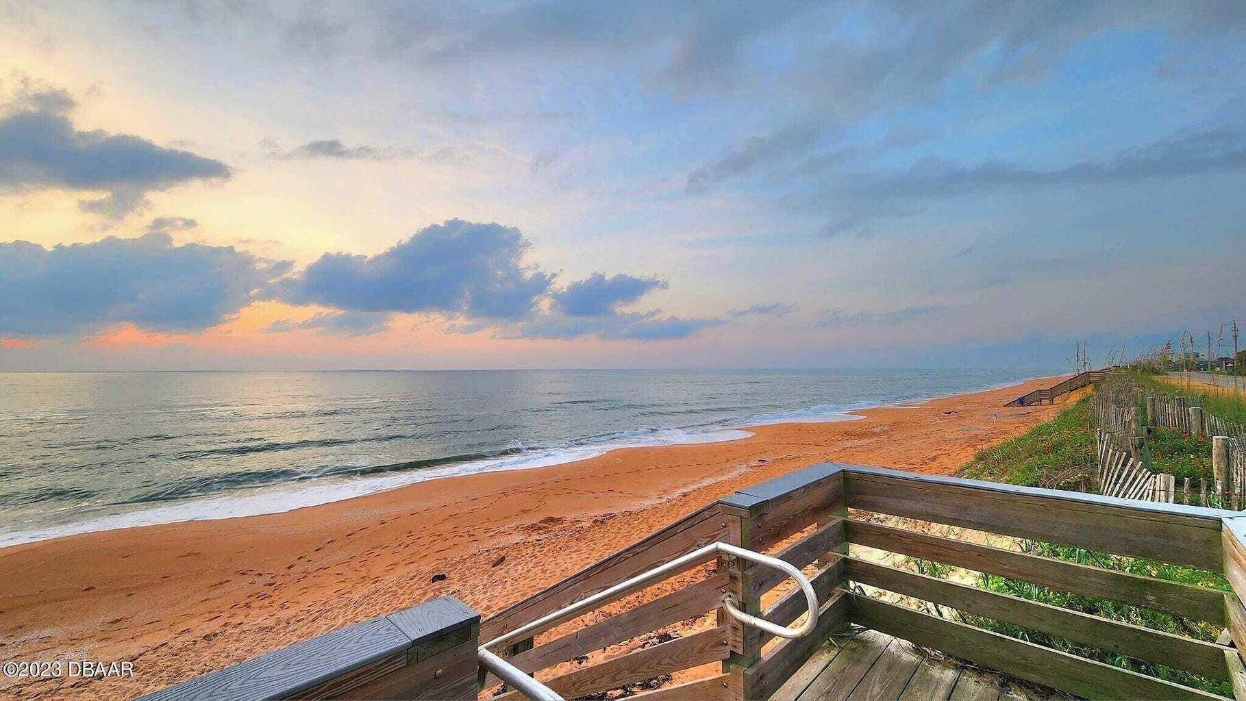 A wooden staircase leading to a beach with a wooden railing and a wooden fence in the background.