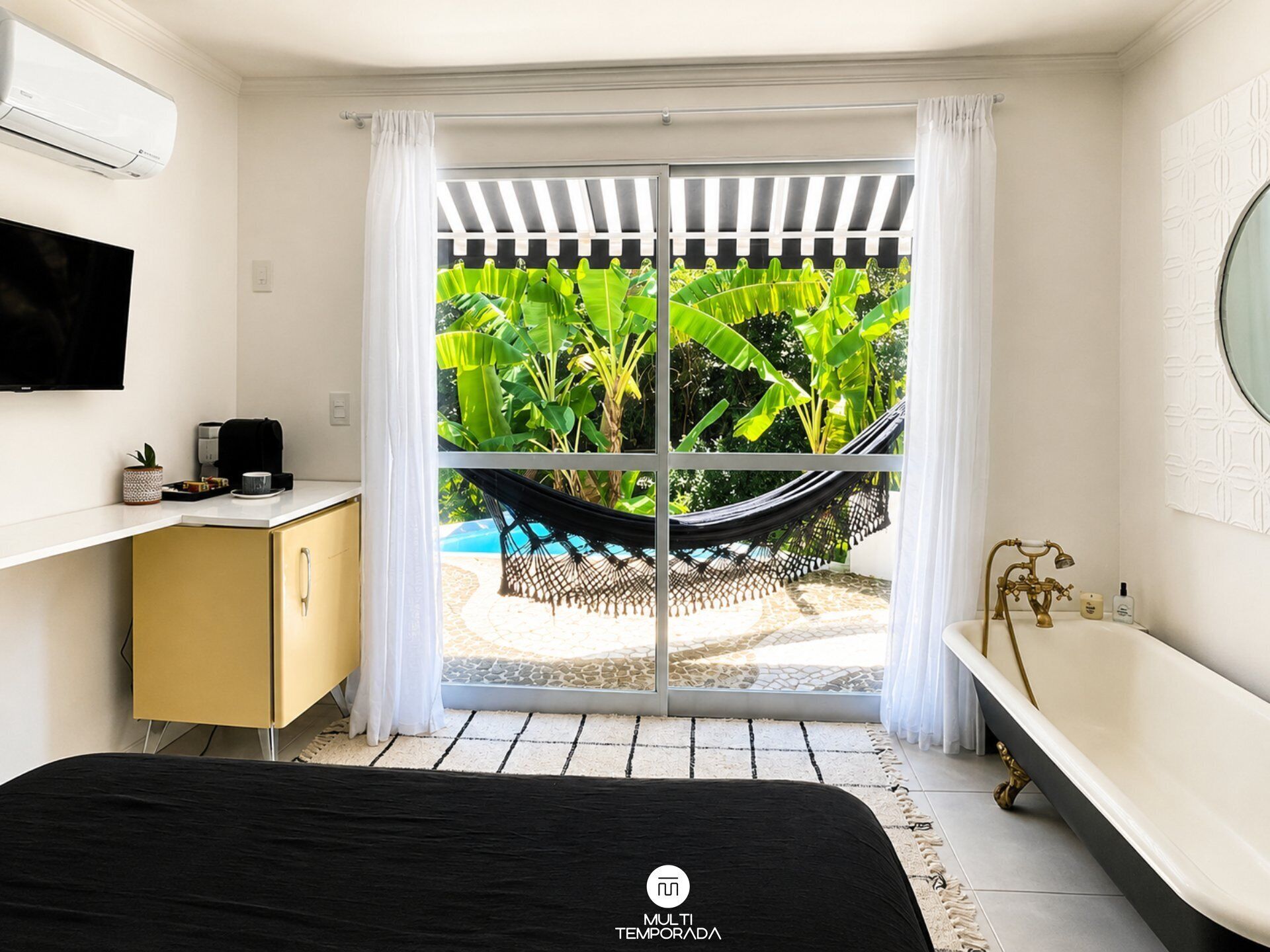 Quarto com cama, escrivaninha, televisão, rede, banheira e vista para piscina e vegetação.