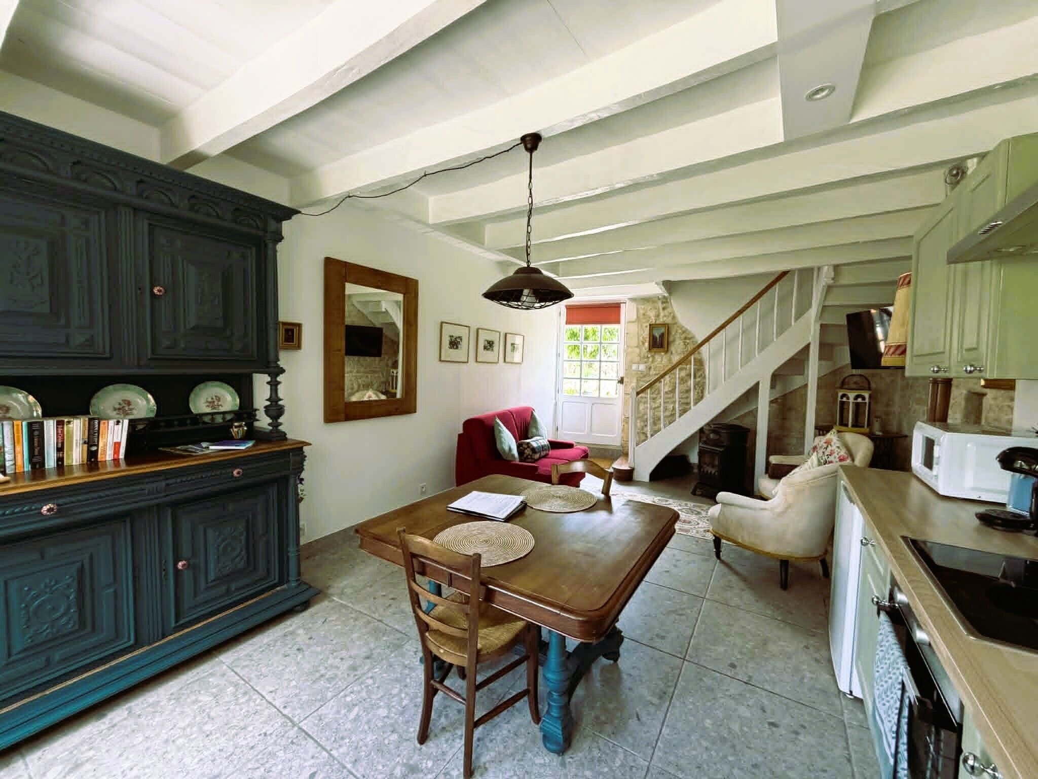 Un comedor con una mesa de madera, sillas y un sofá rojo. Se ve una escalera que sube a otro nivel, y un área de cocina con un microondas.