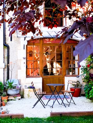 Une cour avec une porte en bois, une petite table et des chaises. On y trouve des plantes en pot et un chemin de gravier.