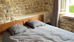 Ein Schlafzimmer mit Steinwand, Holz-Kopfteil, Bett mit grauem Bettzeug, Nachttisch mit Lampe und Fenster mit Vorhängen.