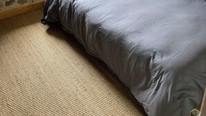 Ein Schlafzimmer mit Steinwand, Holz-Kopfteil, Bett mit grauer Bettwäsche, Fenster mit Vorhängen und Lampe auf dem Nachttisch.