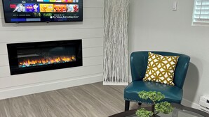 En moderne stue med et fladskærms-tv på væggen, der viser et program, en pejs nedenunder, en turkis sofa med mønstrede puder og et glas sofabord med et bonsaitræ.