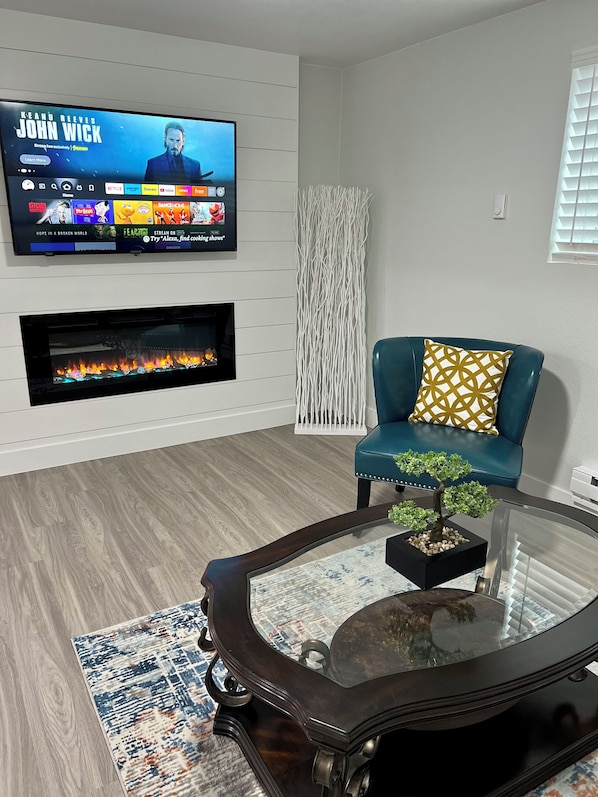 A modern living room with a flat-screen TV mounted on the wall displaying a show, a fireplace below it, a teal sofa with patterned cushions, and a glass coffee table with a bonsai plant.