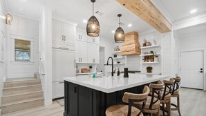 A modern kitchen with white cabinetry, a large island with bar stools, pendant lighting, and a wooden ceiling beam. - 30A Beach House - Dream Chaser by PHG (Santa Rosa Beach)