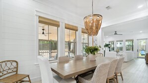 A spacious dining area with a wooden table and wicker chairs, large windows with blinds, and a hanging light fixture. - 30A Beach House - Dream Chaser by PHG (Santa Rosa Beach)