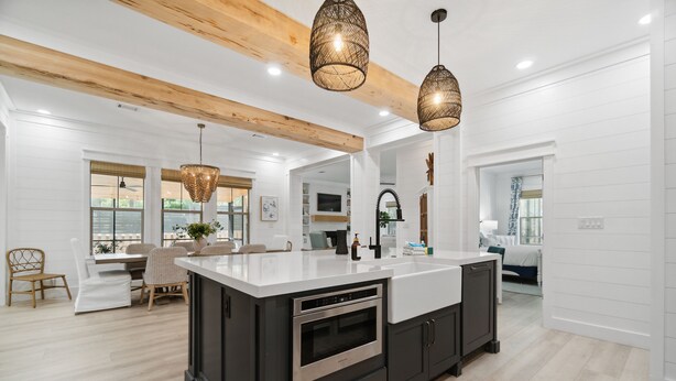 A modern kitchen with a large island, dark cabinetry, and a white countertop. There are hanging pendant lights and a wooden ceiling beam. - 30A Beach House - Dream Chaser by PHG (Santa Rosa Beach)