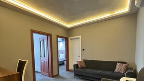 Una sala moderna con un sofá de color oscuro, una mesa de centro de madera y un cojín estampado. Se ve una puerta que da a otra habitación con una cama y una ventana.