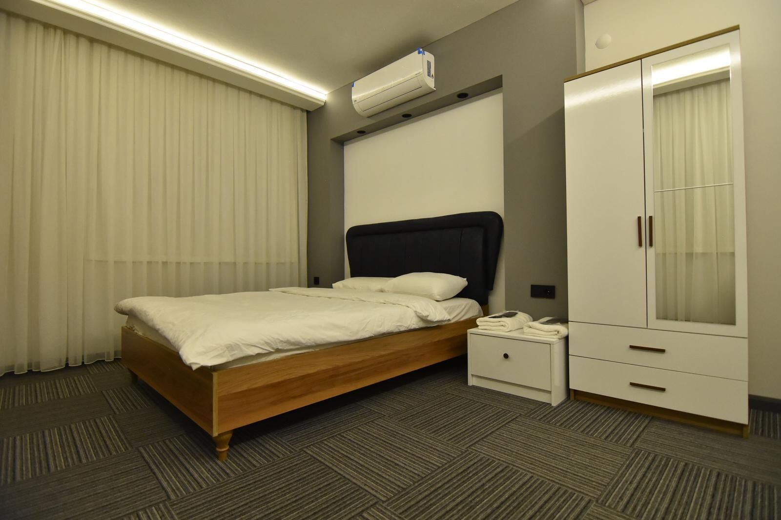 A hotel room with a bed, bedside table, wardrobe, and an air conditioning unit.