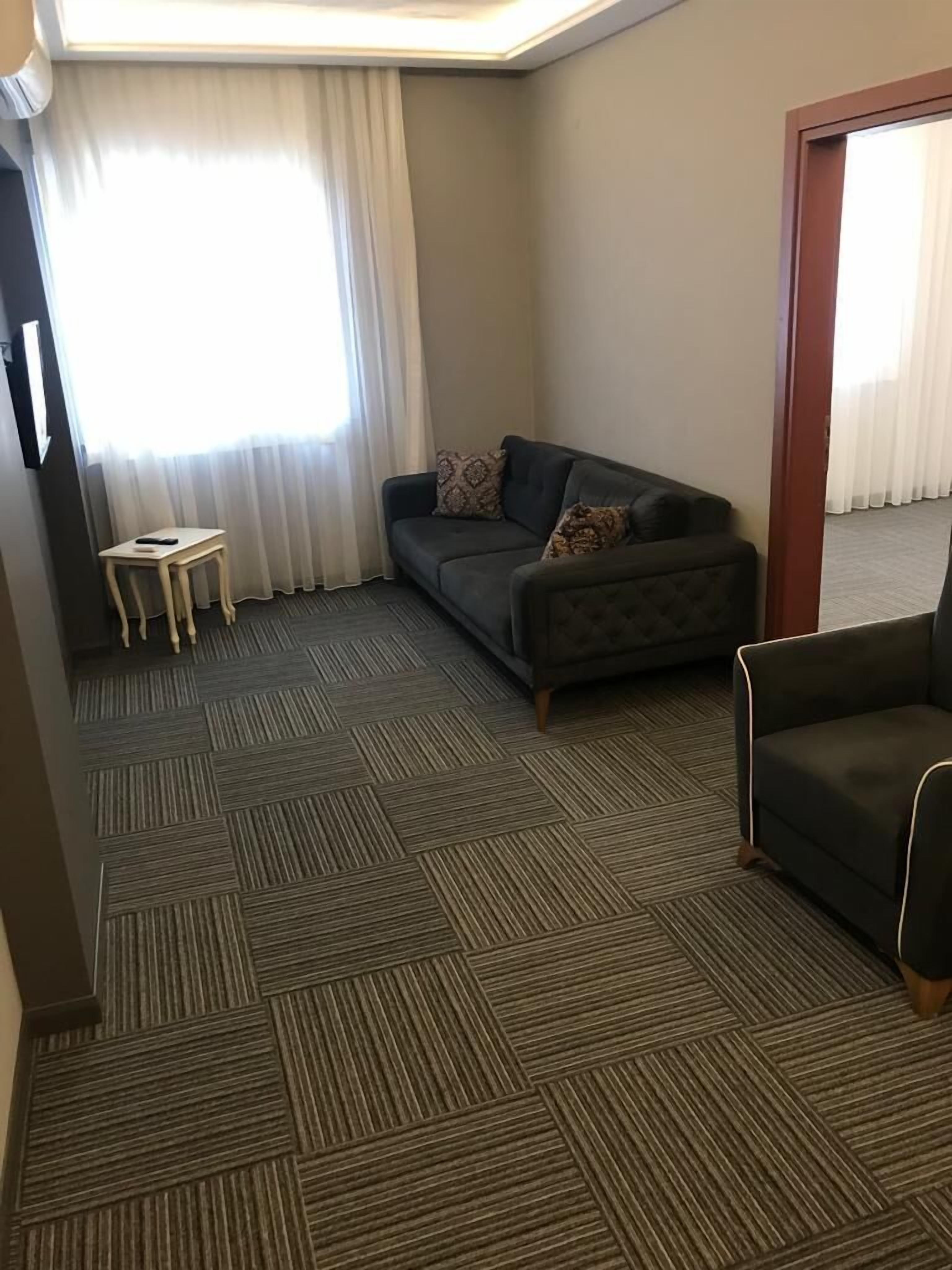 A hotel room with a dark grey sofa, a small wooden side table, and a patterned throw pillow.