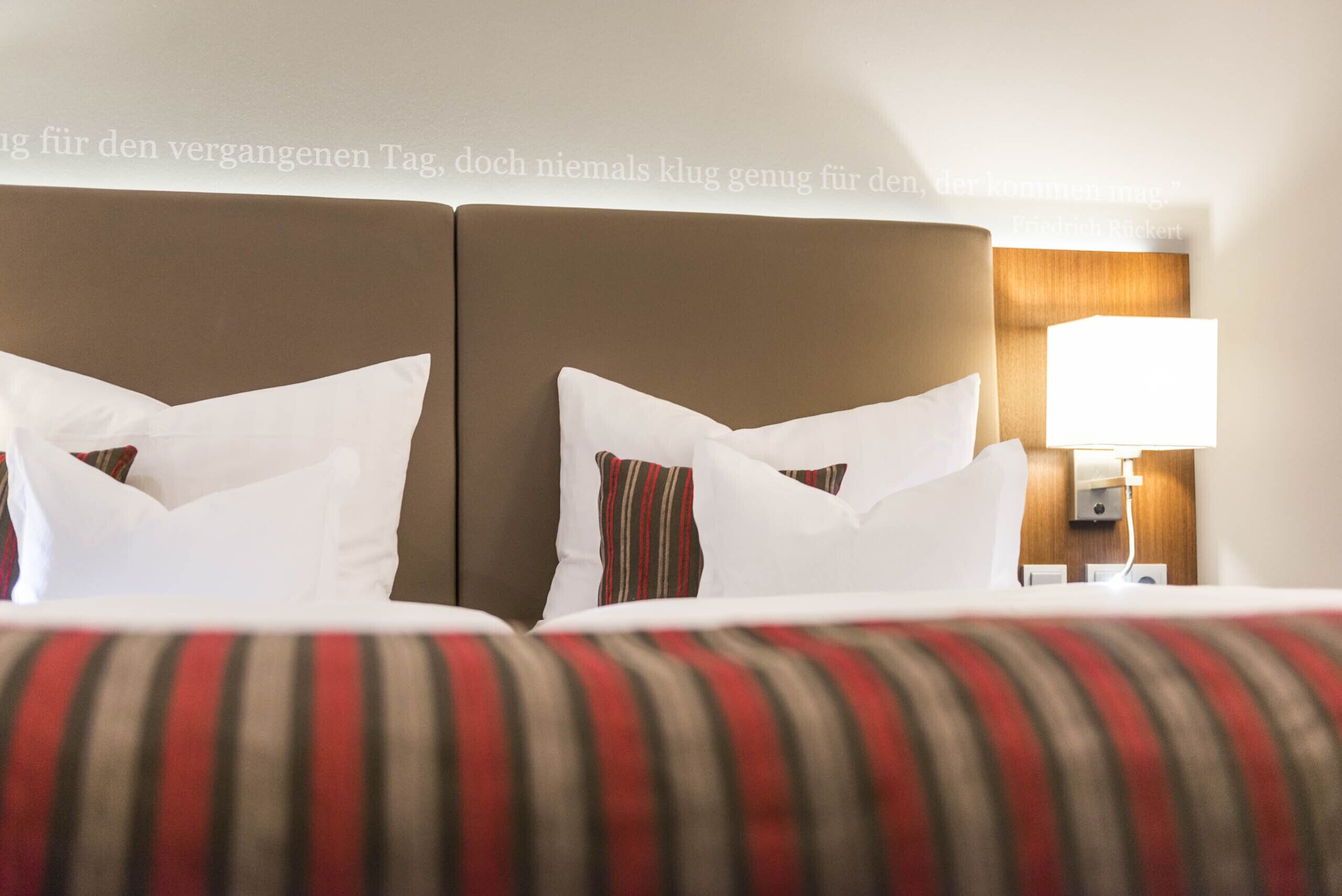 Habitación de hotel con dos camas, cada una con almohadas blancas y a rayas. Se ve un mesita de noche con una lámpara y una pared de paneles de madera.
