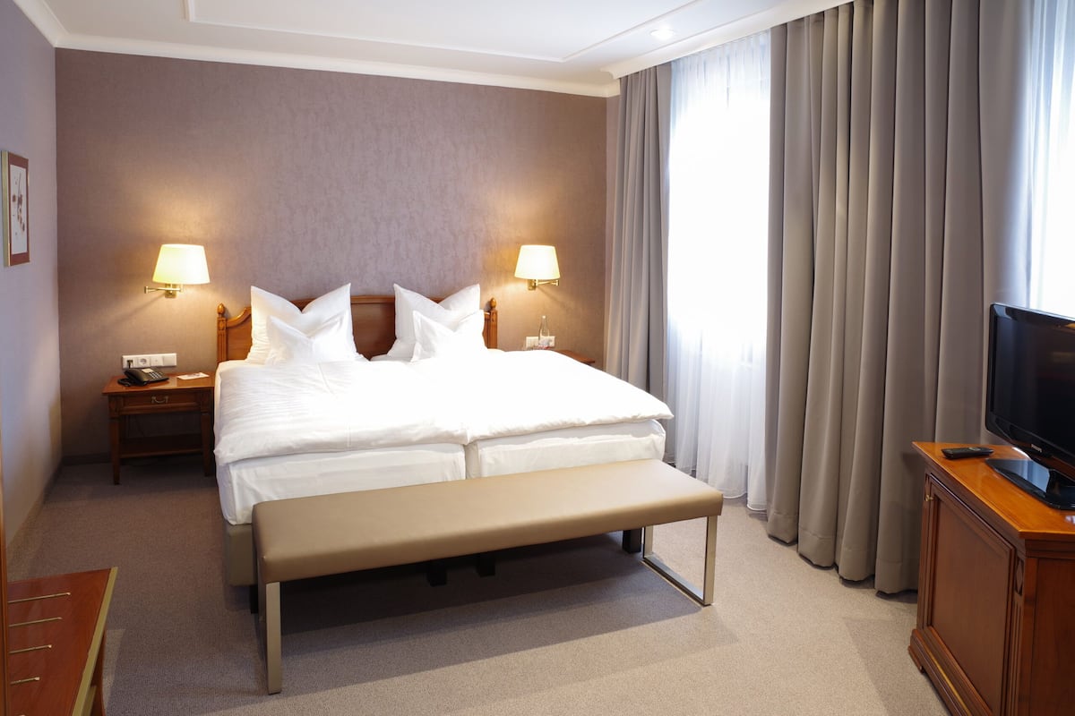 A hotel room with a large bed, two bedside lamps, a bench, a television on a wooden cabinet, and a window with curtains.