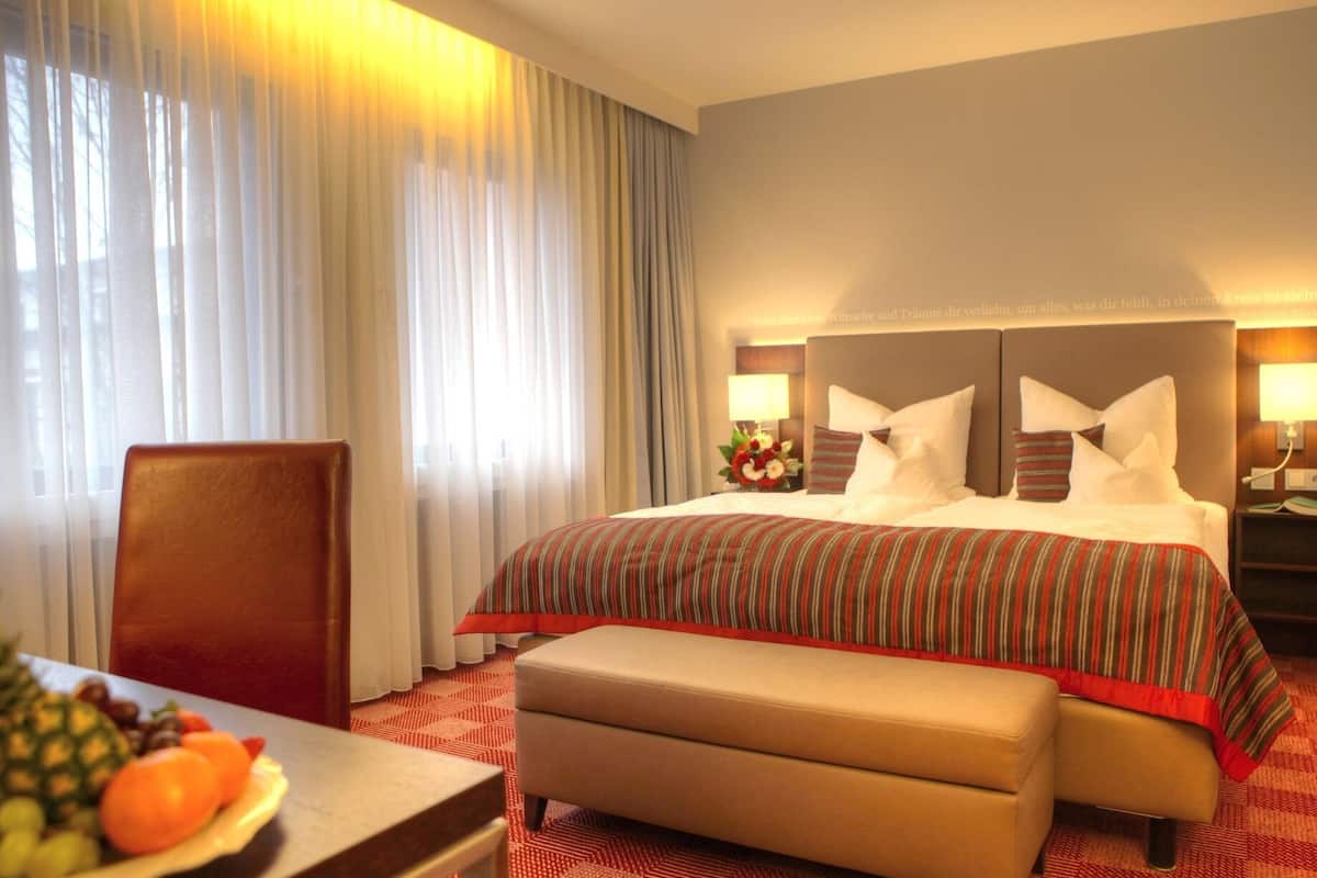 A hotel room with a large bed, a wooden headboard, a bedside table with a lamp, a chair, and a small table with fruit.