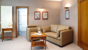 Ein Wohnzimmer mit einem beigen Sofa, einem Holztisch und einem kleinen Beistelltisch. An der Wand hängen zwei gerahmte Bilder und ein Durchgang zu einem anderen Raum.