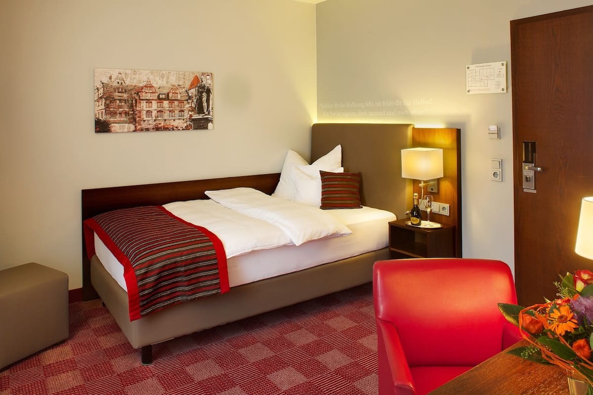A hotel room with a bed, a red armchair, a nightstand with a lamp, and a painting on the wall.