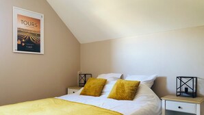 A neatly made bed with white linens and two mustard yellow pillows, a framed poster on the wall, and two bedside tables with black lamps.