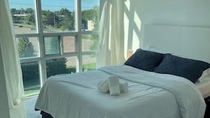A bedroom with a large window, a bed with white bedding, and a view of the outdoors. - Lovely 1 bedroom unit with balcony. Sleeps 1-4 (Whitby)
