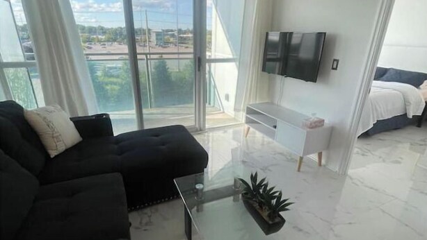 A modern living room with a black sofa, a glass coffee table, and a TV mounted on the wall. - Lovely 1 bedroom unit with balcony. Sleeps 1-4 (Whitby)